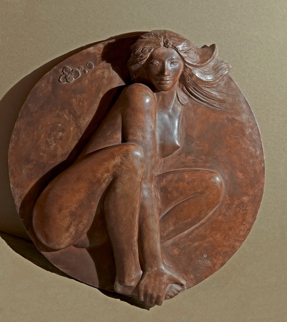Bas-relief en terre cuite, patine bronze, représentant une femme accroupie, ses cheveux flottant derrière elle. Elle tient son pied droit avec ses mains, créant une impression de mouvement et de fluidité dans ses gestes. Cette sculpture s'inscrit dans un cercle.