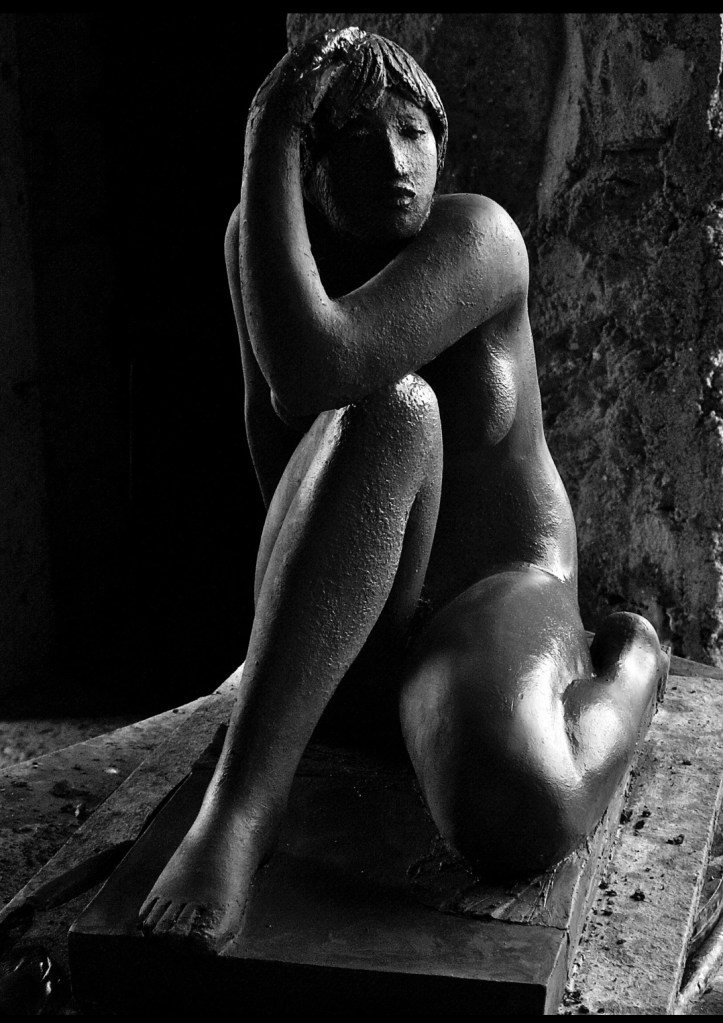 Sculpture en noir et blanc d'une femme assise, nue, avec une posture méditative. Elle tient sa tête avec sa main droite, son genou gauche replié devant elle, créant un jeu d'ombres et de lumières qui souligne les détails texturés de la sculpture.