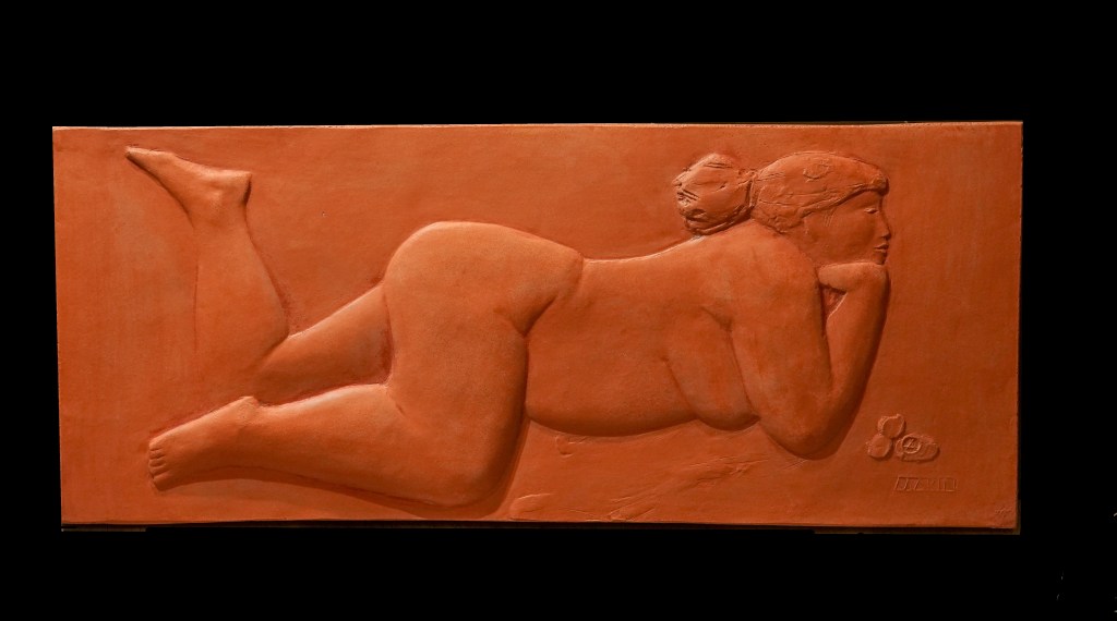 Relief en terre cuite représentant une femme allongée sur le ventre, la tête appuyée sur ses mains et les jambes légèrement pliées. La sculpture met en valeur les formes naturelles du corps avec des détails doux et fluides.