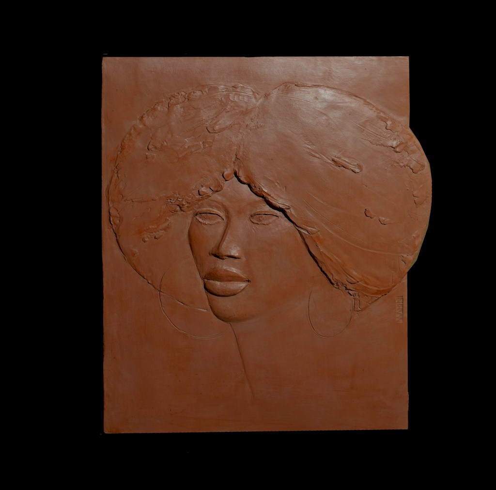Bas-relief représentant un visage féminin avec une coiffure volumineuse qui s'inscrit dans un cercle. Le visage présente des traits élégants, des lèvres pleines, et des boucles d'oreilles rondes visibles sous la coiffure suggérée. La texture de la sculpture accentue le mouvement des cheveux et la douceur des traits du visage.