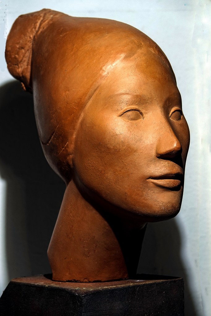 Buste en terre cuite représentant un visage féminin aux traits épurés et une expression sereine. La tête est coiffée d'un chignon, les lignes étirées mettent en valeur la simplicité de la sculpture.